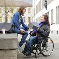 A photograph depicting two young women sitting outside of the Neue Universität and talking to each other. One of the women is sitting in a wheelchair and holding a book.