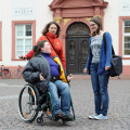 Das Bild zeigt drei junge Frauen, die sich vor dem Gebäude der Alten Universität in Heidelberg unterhalten. Eine der Frauen sitzt in einem Rollstuhl.